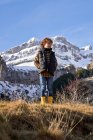 Zufriedenes aktives Kind in warmer Weste und gelben Gummistiefeln mit den Händen in der Tasche, das auf der trockenen Wiese steht und am Fuße der schneebedeckten Berge bei hellem Tag wegblickt — Stockfoto