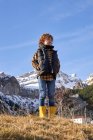 Conteúdo criança ativa em colete quente e botas de borracha amarela com as mãos no bolso em pé no prado seco olhando para longe a pé de montanhas nevadas em dia brilhante — Fotografia de Stock