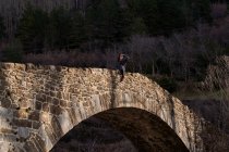 Мандрівна жінка, дивлячись далеко, сидить на старовинному арковому мосту до лісової долини в світлий день — стокове фото