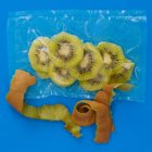 Top view of ripe peeled kiwi in vacuum plastic bag and brown peel on blue background — Stock Photo