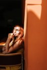 Side view of model in stylish yellow dress with makeup sitting on chair and looking at camera under beam of sun in dark room — Stock Photo