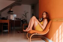 Seitenansicht einer nachdenklichen rothaarigen Frau, die wegschaut, während sie sich zu Hause auf einem Stuhl ausruht und morgens eine Tasse Heißgetränk hält — Stockfoto