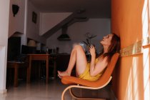 Vista laterale di una donna rossa premurosa che distoglie lo sguardo mentre si riposa sulla sedia a casa con una tazza di bevanda calda al mattino — Foto stock