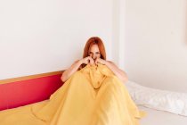 High angle of playful redhead woman sitting and covering half of face with yellow blanket while looking at camera on bed — Stock Photo