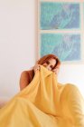 Angle élevé de rousse ludique femme assise et couvrant la moitié du visage avec une couverture jaune tout en regardant la caméra sur le lit — Photo de stock