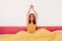 Mujer joven pelirroja satisfecha en vestido amarillo sonriendo mirando a la cámara y cruzando los brazos extendidos despertando en la cama por la mañana - foto de stock