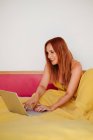 Visão lateral inteligente ruiva freelancer feminino em folhas amarelas trabalhando com laptop deitado na cama — Fotografia de Stock