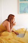 Вид сбоку рыжеволосой женщины-фрилансера в желтых простынях, работающей с ноутбуком, лежащим на кровати — стоковое фото