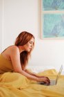 Side view of redhead female freelancer in yellow sheets working with laptop lying on bed — Stock Photo