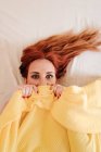 Draufsicht der überraschten rothaarigen lustigen Frau, die lächelt, während sie zu Hause unter einer gelben Decke hervorblickt — Stockfoto