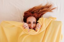 Top view of surprised redhead funny woman smiling while looking out from under yellow blanket at home — Stock Photo