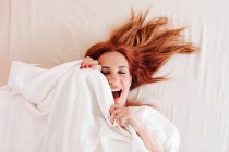 De cima vista superior da mulher engraçada ruiva surpresa sorrindo enquanto olha para fora de baixo cobertor branco em casa — Fotografia de Stock