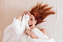 Сверху вид удивленной рыжеволосой забавной женщины, улыбающейся, глядя из-под белого одеяла дома — стоковое фото