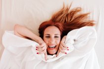 Von oben Ansicht der überraschten rothaarigen lustigen Frau, die lächelt, während sie unter einer weißen Decke nach Hause schaut — Stockfoto