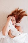 Сверху вид удивленной рыжеволосой забавной женщины, улыбающейся, глядя из-под белого одеяла дома — стоковое фото