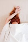 Vista superior del pelo rojo de la señora sin rostro con el brazo sobresaliente que tiene relajación mientras está acostado en la cama bajo sábanas blancas - foto de stock