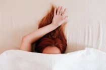 Visão superior do cabelo vermelho da senhora sem rosto com braço saliente ter relaxamento enquanto deitado na cama sob lençóis brancos — Fotografia de Stock