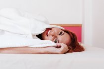 Cheerful redhead funny woman smiling while looking out from under white blanket at home — Stock Photo