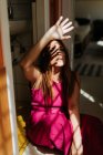 Продумана руда жінка в стильній рожевій сукні сидить з схрещеними ногами на підлозі і дивиться в сторону з променями сонця на обличчі — стокове фото