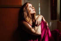 Thoughtful redhead female in stylish pink gown sitting with crossed legs on floor and looking away with rays of sun on face — Stock Photo