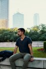 Junge hispanische männliche Studenten in stylischer Freizeitkleidung sitzen auf einem Bauzaun und schauen mit Blick auf die Innenstadt im Hintergrund in Dallas, Texas — Stockfoto