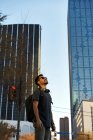 Homem hispânico barbudo em roupa casual sorrindo e olhando para cima com óculos de sol na moda e fones de ouvido no pescoço na rua da cidade — Fotografia de Stock