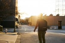 Вид ззаду на невпізнаваного чоловіка в повсякденному вбранні з рюкзаком, що йде по міській вулиці під час заходу сонця — стокове фото