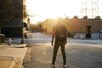 Обратный вид на неузнаваемого мужчину в повседневной одежде с рюкзаком, гуляющего по городской улице во время заката — стоковое фото