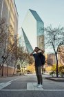 Viajante masculino hispânico em roupa casual e óculos de sol com mochila colocando fones de ouvido enquanto passeia no centro da cidade — Fotografia de Stock