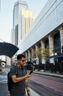 Vue latérale d'un touriste hispanique contemporain en tenue décontractée et lunettes de soleil avec sac à dos et écouteurs utilisant le téléphone portable dans la rue moderne de la ville — Photo de stock