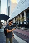 Вид сбоку современного латиноамериканского туриста в повседневной одежде и солнцезащитных очках с рюкзаком и наушниками с помощью мобильного телефона на современной городской улице — стоковое фото