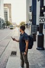 Homem hispânico anônimo em roupas casuais com mochila surfando no celular enquanto estava na plataforma na rua da cidade — Fotografia de Stock