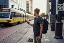 Anonymer hispanischer Mann in Freizeitkleidung mit Rucksack surft auf Handy, während er auf Bahnsteig an der Stadtstraße steht — Stockfoto