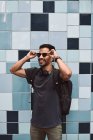 Hombre hispano feliz en ropa casual y mochila con auriculares de pie escuchando música en la calle de la ciudad - foto de stock