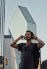 Glücklicher hispanischer Mann mit geschlossenen Augen in Freizeitkleidung und Rucksack mit Kopfhörern, der an der Stadtstraße Musik hört — Stockfoto