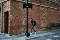 Іспанський чоловік учень у повсякденному одязі з рюкзаком, що йде по порожній вулиці міста з цегляним будинком на задньому плані. — стокове фото