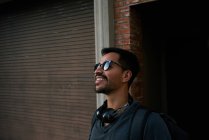 Vista lateral do viajante masculino hispânico em roupa casual e óculos de sol estilista com mochila de pé ao longo da rua da cidade vazia com edifício de tijolos no fundo — Fotografia de Stock