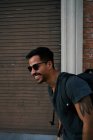 Vista lateral do viajante masculino hispânico em roupa casual e óculos de sol estilista com mochila de pé ao longo da rua da cidade vazia com edifício de tijolos no fundo — Fotografia de Stock