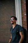 Vista lateral del viajero masculino hispano en traje casual y gafas de sol estilista con mochila de pie a lo largo de la calle vacía de la ciudad con edificio de ladrillo en el fondo - foto de stock