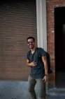 Hombre hispano viajero en traje casual y gafas de sol estilista con mochila de pie a lo largo de la calle vacía de la ciudad con el edificio de ladrillo en el fondo - foto de stock