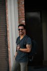 Viaggiatore maschio ispanico in abito casual e occhiali da sole stilista con zaino in piedi lungo la strada vuota della città con edificio in mattoni sullo sfondo — Foto stock