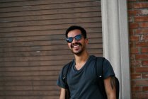 Латиноамериканец в повседневной одежде и стилистские солнцезащитные очки с рюкзаком, стоящим вдоль пустой городской улицы с кирпичным зданием на заднем плане — стоковое фото