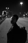 Vue latérale de l'homme hispanique à la mode en veste décontractée en cuir noir avec sac à dos regardant la caméra avec la ville de nuit sur fond flou — Photo de stock