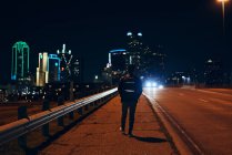 Вид ззаду анонімного чоловіка в повсякденній чорній шкіряній куртці з рюкзаком, що йде в нічному місті на розмитому фоні — стокове фото