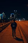 Visão traseira do homem anônimo com jaqueta de couro preto casual com mochila andando na cidade noturna em fundo embaçado — Fotografia de Stock