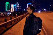 Вид сбоку на модного латиноамериканца в обычной черной кожаной куртке с рюкзаком, смотрящего в сторону с ночным городом на размытом фоне — стоковое фото