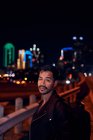 Vista lateral del hombre hispano de moda en chaqueta de cuero negro casual con mochila mirando a la cámara con la ciudad de la noche sobre fondo borroso - foto de stock