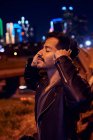 Задоволений латиноамериканець в стильній шкіряній куртці посміхається, слухаючи музику з навушниками на сучасній вулиці — стокове фото