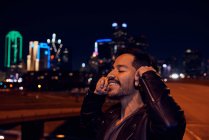 Zufriedener hispanischer Mann in stylischer schwarzer Lederjacke lächelt, während er in der modernen Stadtstraße Musik mit Kopfhörern hört — Stockfoto