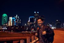 Modischer hispanischer Mann in lässiger schwarzer Lederjacke mit Rucksack, der auf verschwommenem Hintergrund in die Kamera mit nächtlicher Stadt blickt — Stockfoto